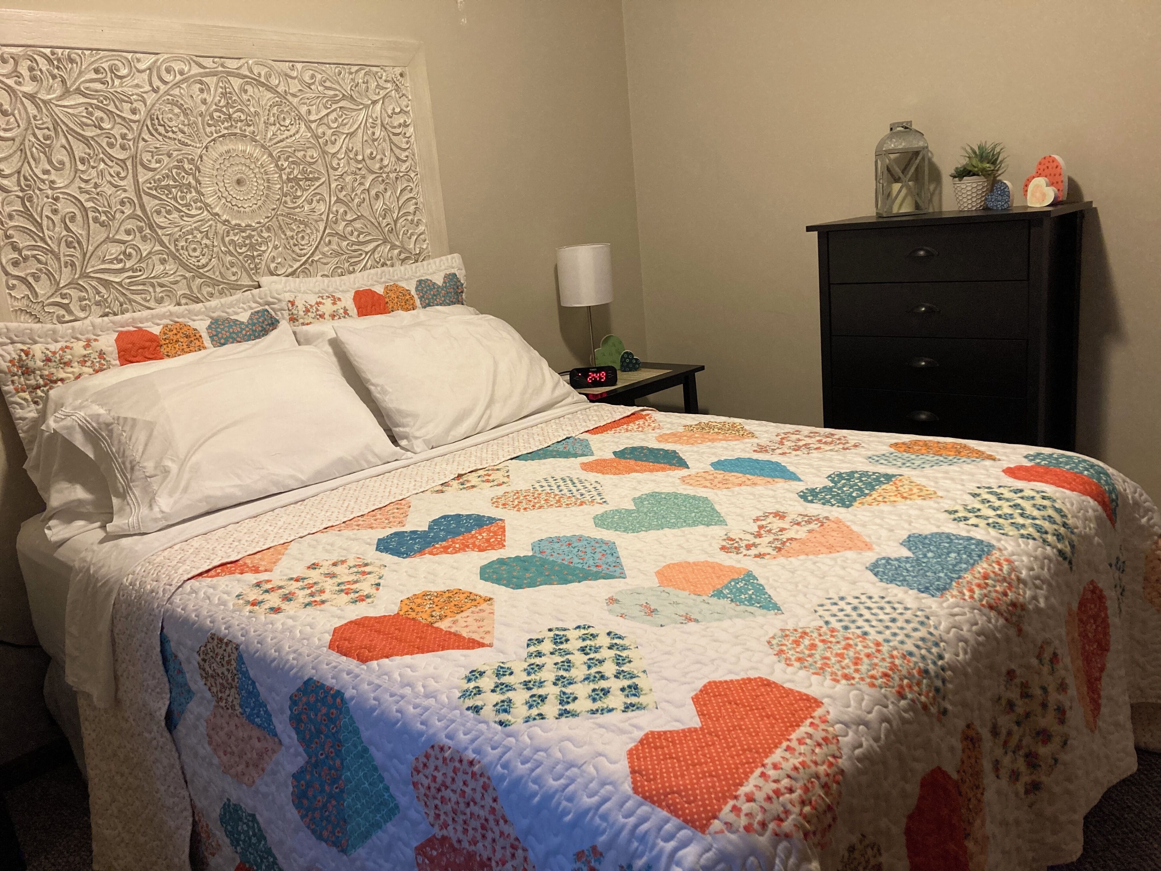 Una cama bien hecha con un colcha colorida, un cabecero decorativo, una mesita de noche con una lámpara y una pequeña mesita con un reloj.