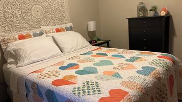 Una cama bien hecha con un colcha colorida, un cabecero decorativo, una mesita de noche con una lámpara y una pequeña mesita con un reloj.