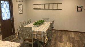 Zona pranzo con tovaglia a scacchi, sei sedie, una pianta in vaso e una mensola in legno al muro.