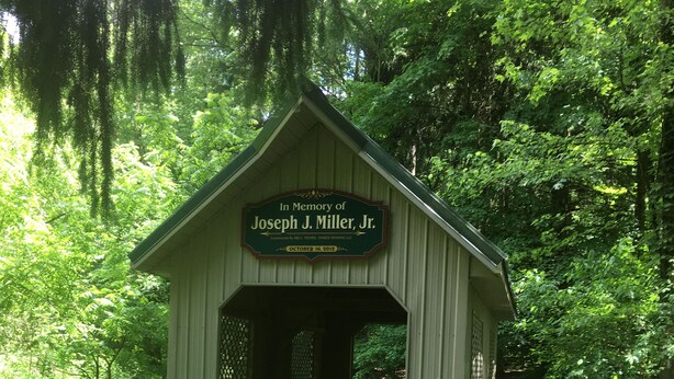 Un ponte coperto in legno con una targa commemorativa dedicata a Joseph J. Miller, Jr., immerso in una vegetazione lussureggiante.