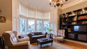 Un soggiorno con un divano in pelle marrone, una poltrona beige, un poggiapiedi nero e una libreria piena di libri e oggetti decorativi.