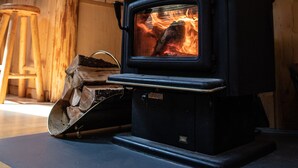 A wood-burning stove with a chimney, logs, and a metal grate in a wooden interior. - Pine Lodge - 9 beds;  Extra fees  for 7+ Hot Tub Sauna AC EV Charger Pets (Packwood)