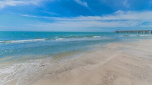 Een strand met helder water, een steiger die in de zee uitloopt, en een heldere lucht.