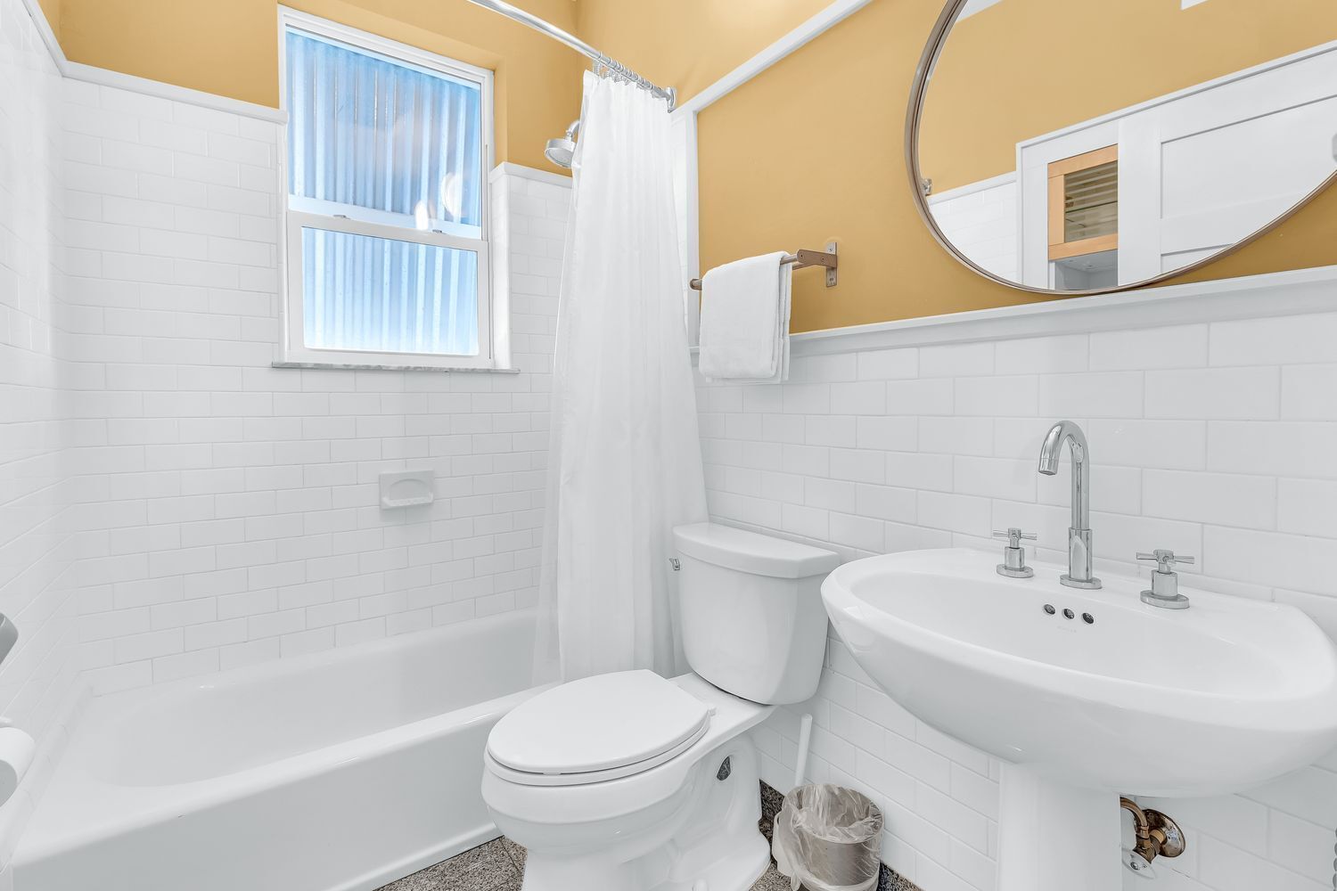 Une salle de bain équipée d’une baignoire, de toilettes et d’un lavabo, avec des carreaux blancs et une fenêtre avec des stores.