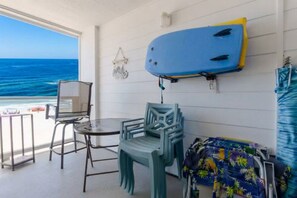 A room with a view of the ocean, a blue surfboard mounted on the wall, a small table with chairs, and a colorful beach towel.