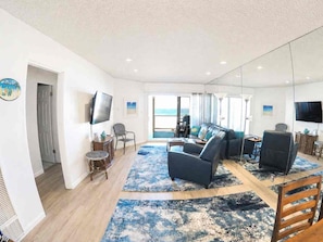 A modern living room with a large TV, comfortable seating, a view of the ocean, and a large mirror.