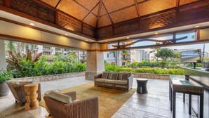 Un espace intérieur vaste et lumineux, avec un plafond haut, des meubles en osier et une vue sur un jardin luxuriant et des bâtiments.