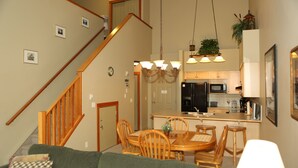A dining area with a wooden table and chairs, a chandelier, and a kitchen in the background.
