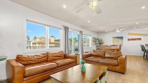 A spacious living room with leather sofas, a wooden coffee table, and large windows offering a view of buildings and palm trees.