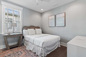 Quarto com cama, mesa de cabeceira, abajur, janela com persianas e duas imagens emolduradas na parede.