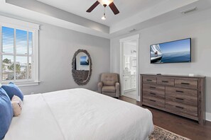 Un dormitorio con una cama grande, un televisor montado en la pared, un ventilador de techo y vistas al mar a través de la ventana.