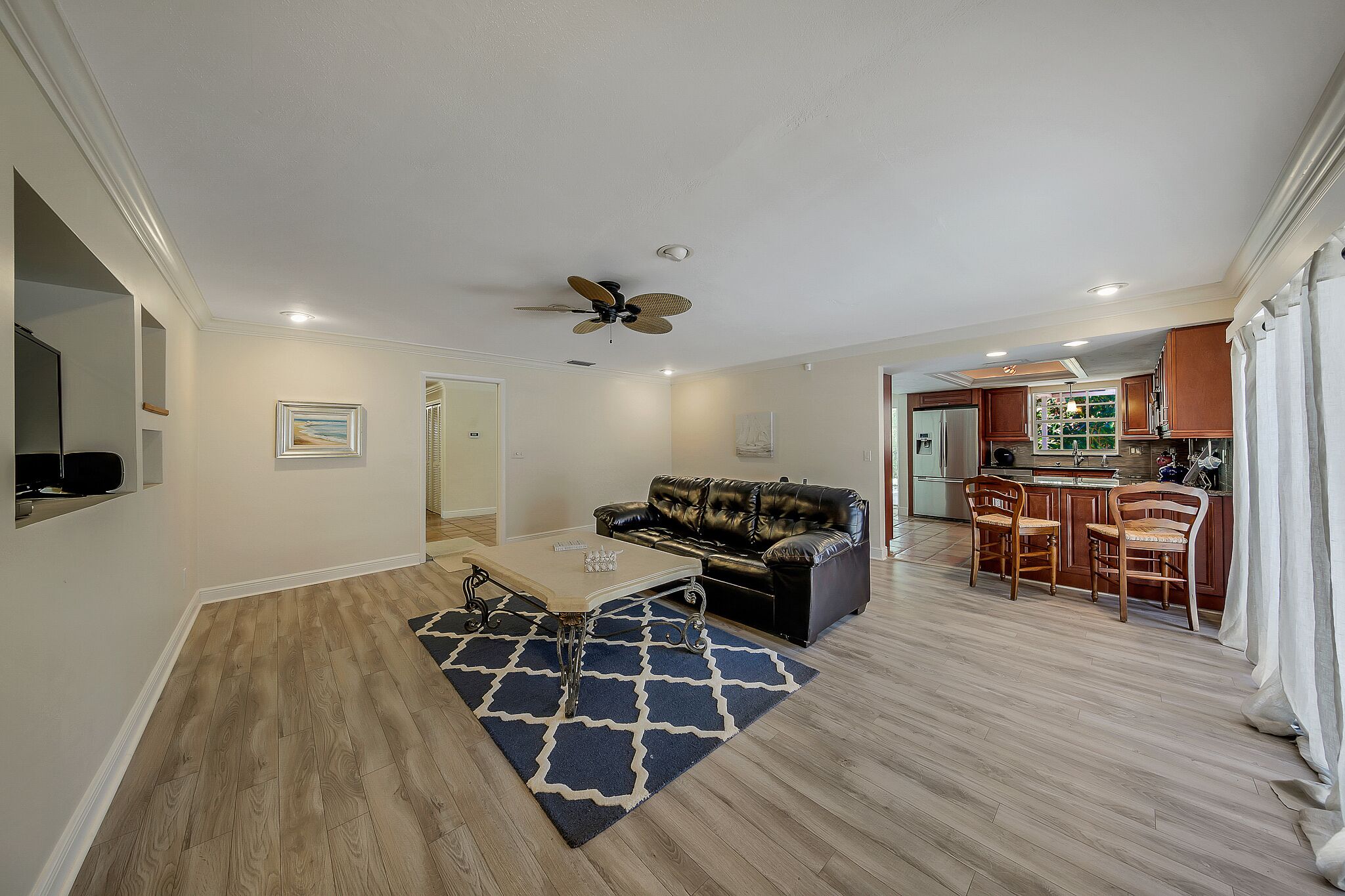 A living room with a sofa, coffee table, and a ceiling fan. There is a kitchen area with wooden cabinets and a dining set.
