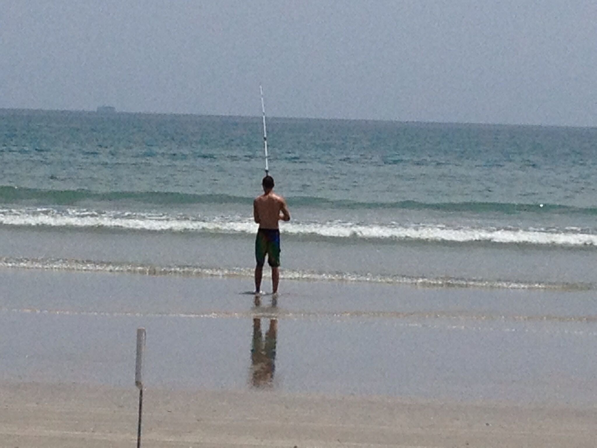 A person standing on a beach with a fishing rod, facing the ocean.