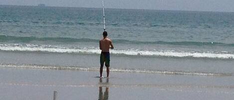 A person standing on a beach with a fishing rod, facing the ocean.
