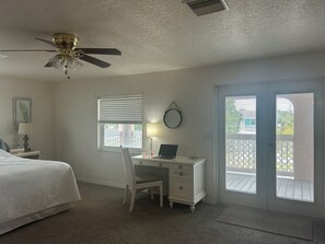 A bedroom with a bed, a desk with a laptop, a chair, a ceiling fan, a window with blinds, and a door leading to an outdoor area.