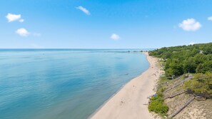A coastal beach with a clear view of the sea, a sandy shoreline, and a forested hillside with a staircase leading down to the beach. - Luxury Macatawa Beach Home with Stunning Lake Michigan Views (Holland)