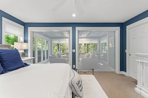 A bedroom with a bed, a nightstand, a lamp, and a view of a balcony with outdoor furniture. - Luxury Macatawa Beach Home with Stunning Lake Michigan Views (Holland)