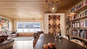 Ein modernes Wohnzimmer mit Holzbalkendecke, einem großen Fenster, einer Couch, einem Esstisch mit Früchten und Weingläsern sowie einem Bücherregal.