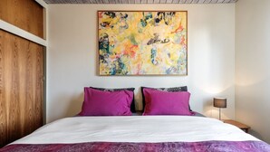 Ein Schlafzimmer mit einem großen Bett, zwei violetten Kissen, einem farbenfrohen abstrakten Gemälde an der Wand und einer Holzvertäfelung links.