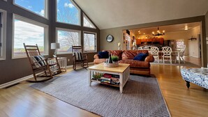 Amplia sala de estar con ventanas grandes, una mesa de centro de madera, un sofá de cuero marrón y un comedor con sillas blancas.