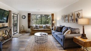 Un salon avec un canapé, une table basse et un téléviseur. Il y a des fenêtres, une horloge et des œuvres d’art accrochées aux murs.