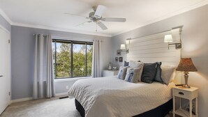 Une chambre à coucher comprenant un lit, une table de chevet, une lampe, un ventilateur de plafond, une fenêtre avec des rideaux et une vue sur les arbres à l’extérieur.