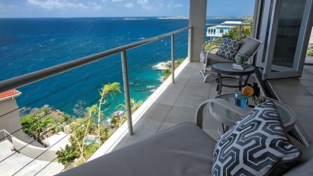 A balcony with a view of the ocean, a table with chairs, and a patterned cushion.