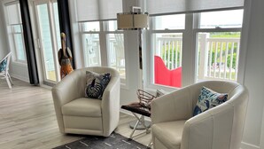 A modern living room with two white armchairs, a glass coffee table, and a large window with a view of a balcony. - Rest, Relax & Enjoy Your Stunning Water Views in the Heart of Southport, NC! (Southport)