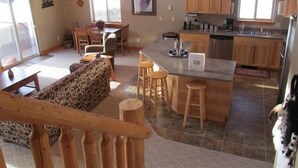 Un vaste espace de vie comprenant un banc en bois, un canapé et une table à manger avec des chaises. On aperçoit également une cuisine avec des meubles en bois et un coin bar.