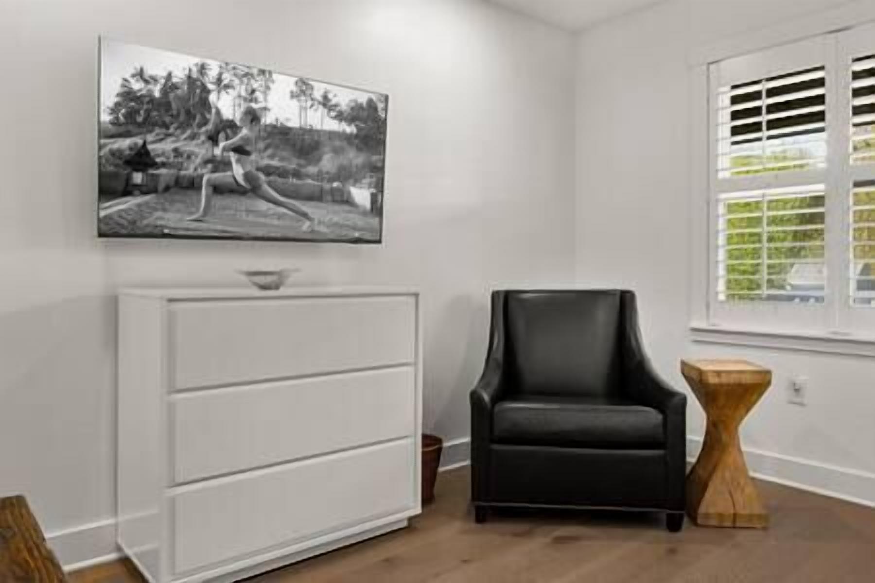 A room with a white dresser, a black leather chair, a wooden side table, and a window with blinds.