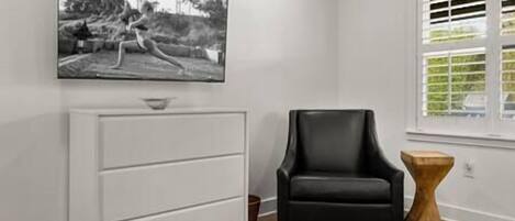 A room with a white dresser, a black leather chair, a wooden side table, and a window with blinds.