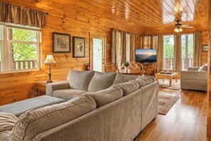 A cozy cabin interior with a sectional sofa, a television, wooden floors, and large windows. - 6 BD 6 BATH ALL Ensuite cabin. Come and make memories for a lifetime! (Sevierville)