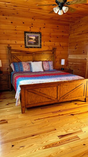 Een slaapkamer in een houten blokhut, met een bed, nachtkastjes, een plafondventilator en een lijst met een foto aan de muur.