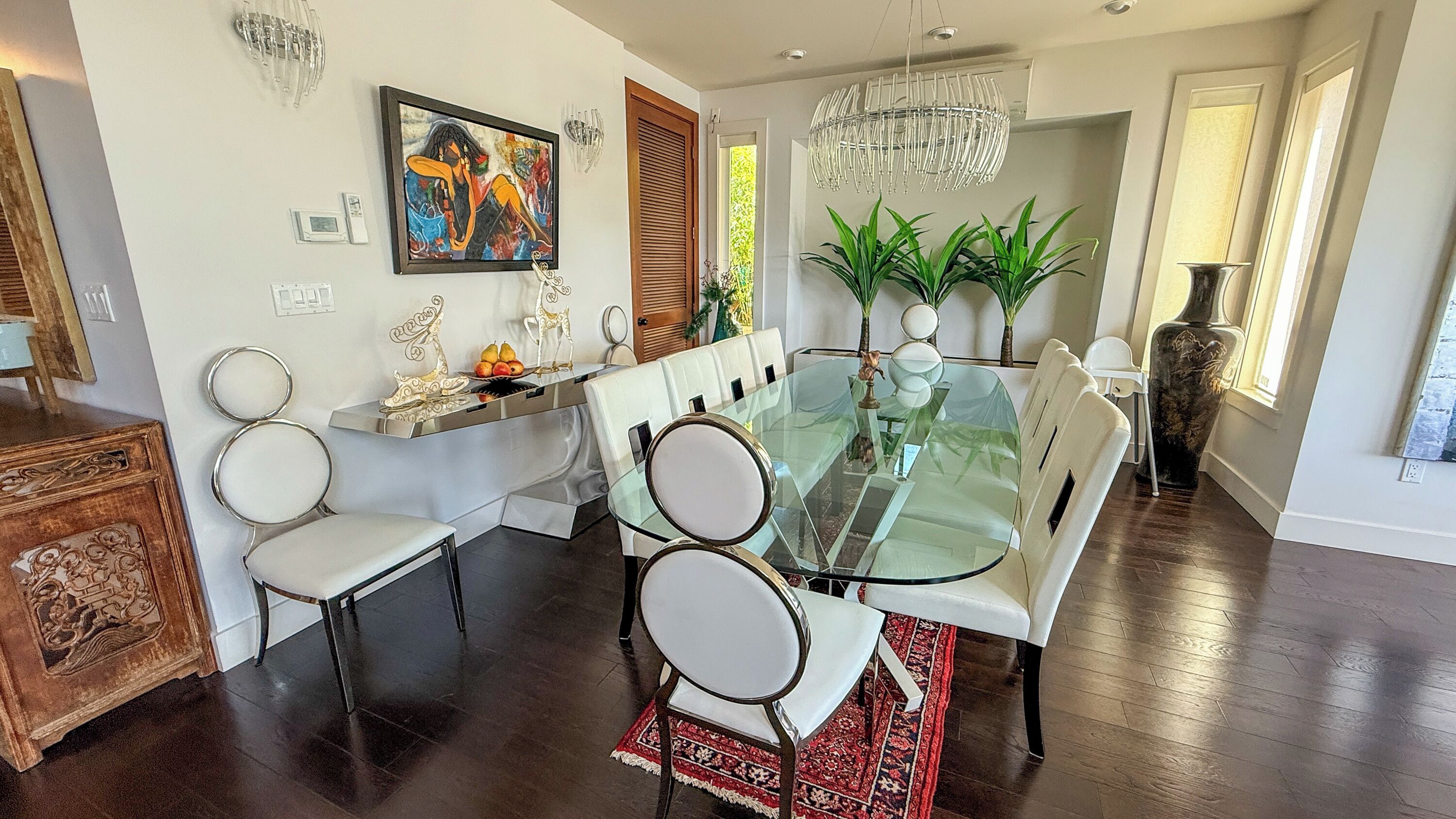 A dining area with a glass table, white chairs, a chandelier, and a painting on the wall.