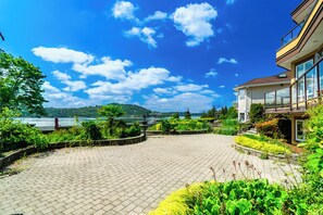 A paved courtyard with a view of a lake and mountains, surrounded by lush greenery and a modern building with large windows.