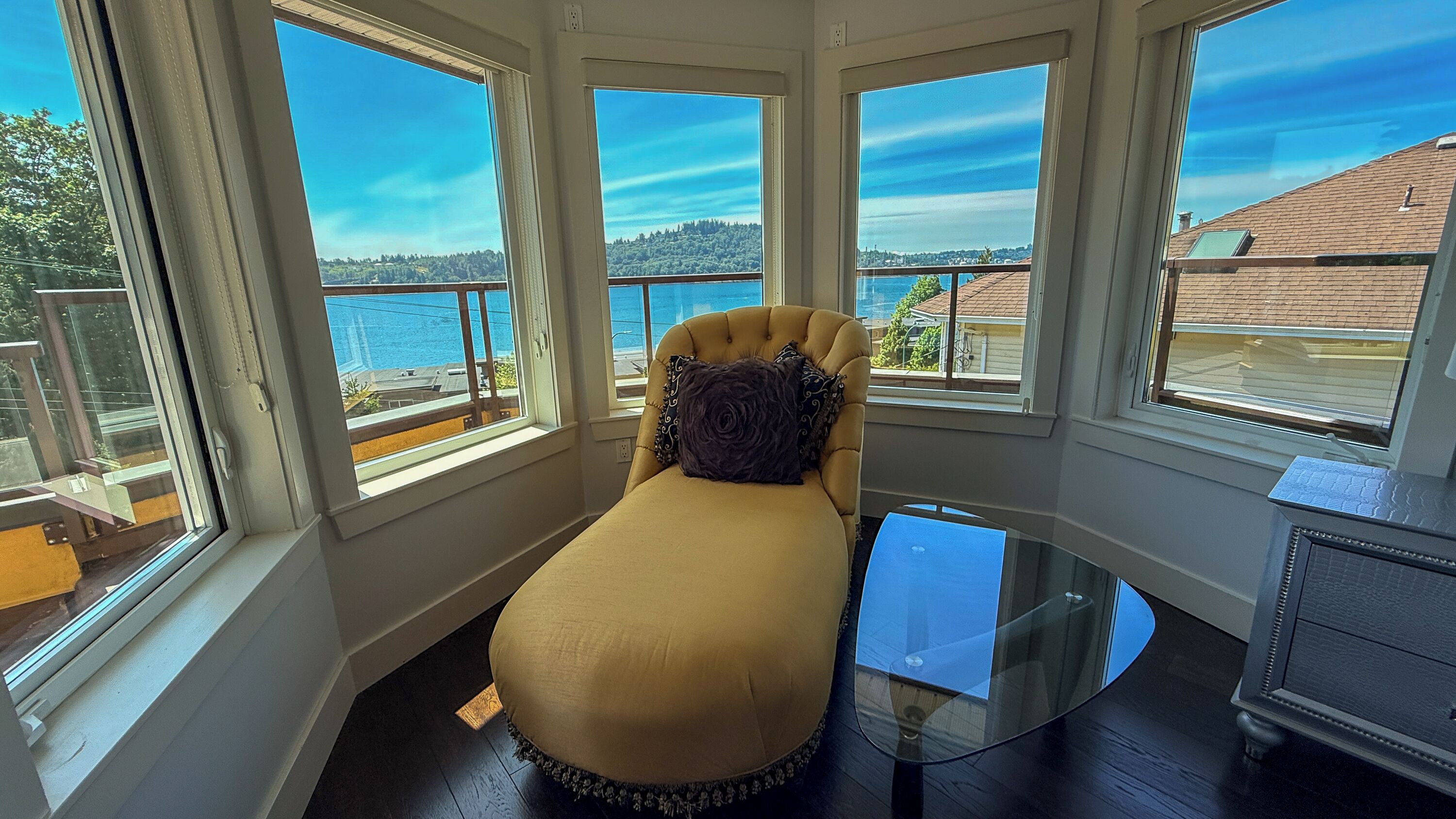 A room with a bed, a glass table, and large windows with a view of a body of water and hills.