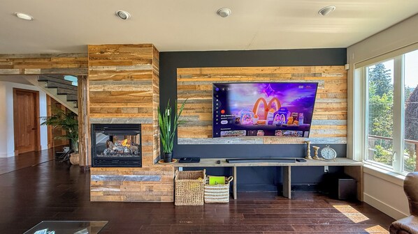 A modern living room with a fireplace, a large flat-screen TV mounted on a wooden wall, and a view of the outdoors through a large window.