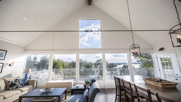 A spacious room with a high ceiling, large windows offering a view of a deck and trees, a dining area with wooden chairs and a table, and a hanging light fixture. - Perfect Location For Your Lake Getaway (Cadiz)