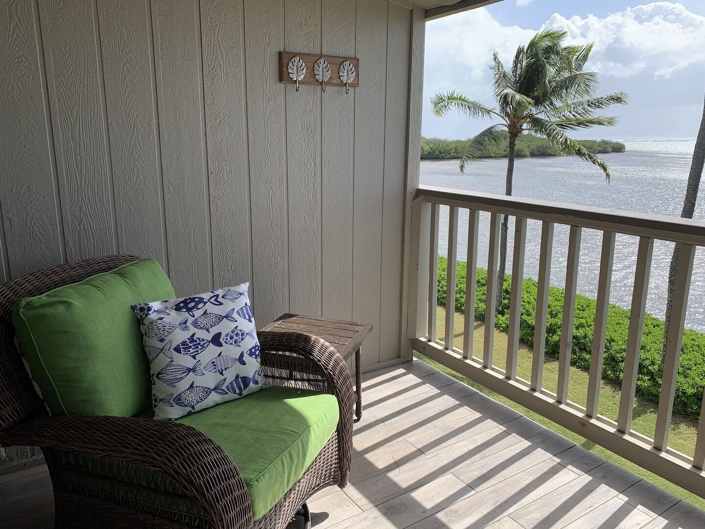 A wicker chair with a green cushion and a blue fish-patterned pillow, a small wooden table, and a view of a palm tree and water through a balcony railing.
