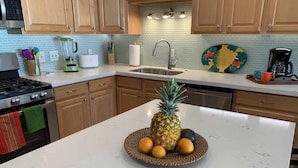 A kitchen with wooden cabinets, a white marble countertop, a stainless steel sink, and a fruit bowl with a pineapple and oranges.