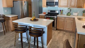 A modern kitchen with wooden cabinets, stainless steel appliances, a central island with bar stools, and a tiled backsplash.