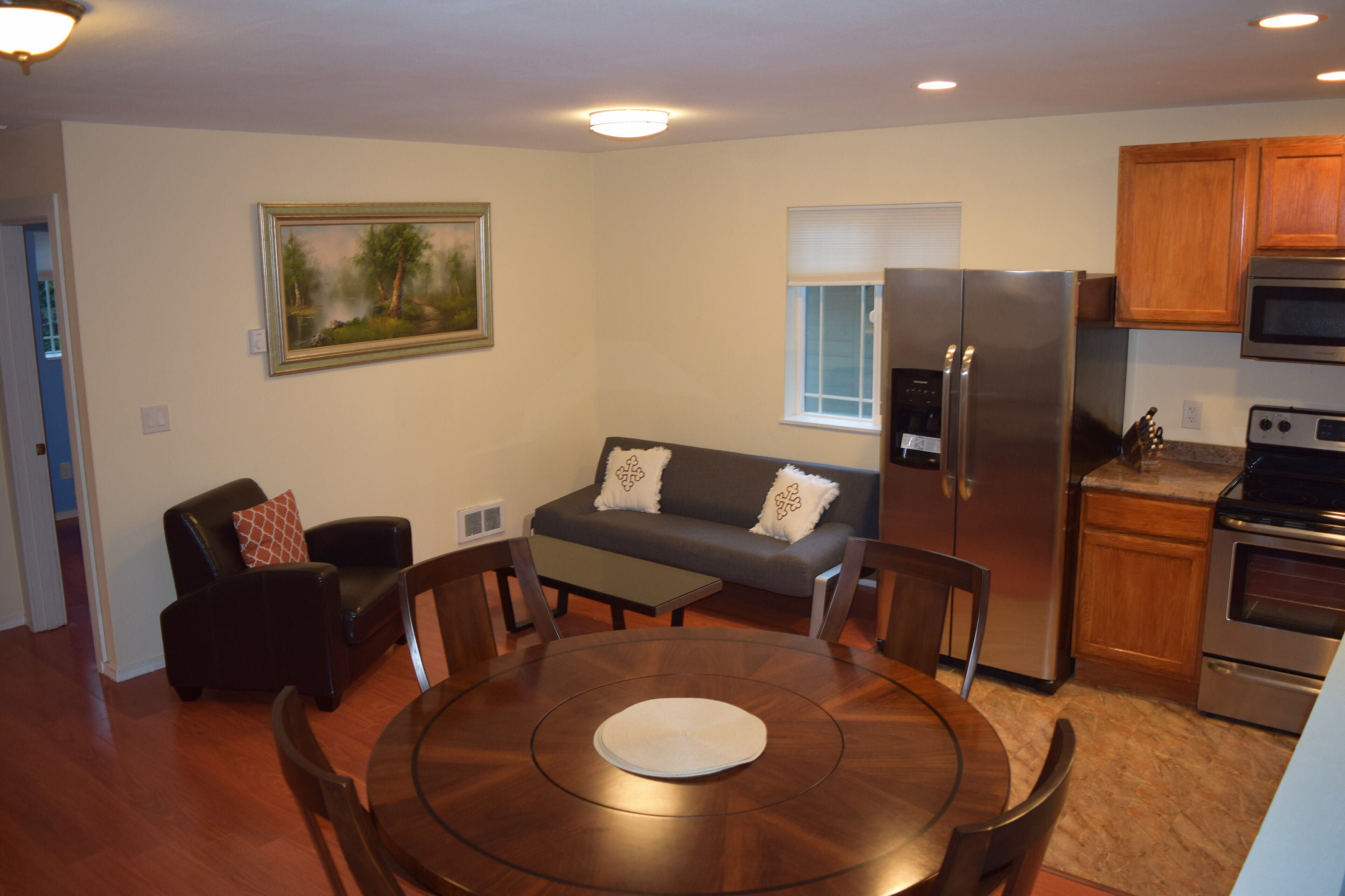 A modern kitchen with a refrigerator, oven, and microwave. There is a dining area with a round table and chairs. A sofa and a chair are also visible.