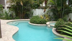 Una piscina curva con borde de piedra, rodeada de exuberante vegetación y una valla blanca.