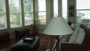 Sala de estar con sofá, una mesita de centro y una lámpara. Amplias ventanas con persianas, una planta en maceta y vistas al exterior.