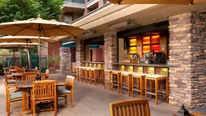 An outdoor patio area with wooden tables and chairs, stone pillars, and a bar with illuminated shelves.