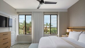 A hotel room with a bed, a television, a ceiling fan, a window with a view of palm trees, and a nightstand with a lamp. - Confirmed Reservation - March 13–20, 2027 - 1BR Premium Villa - Instant Book (Scottsdale)