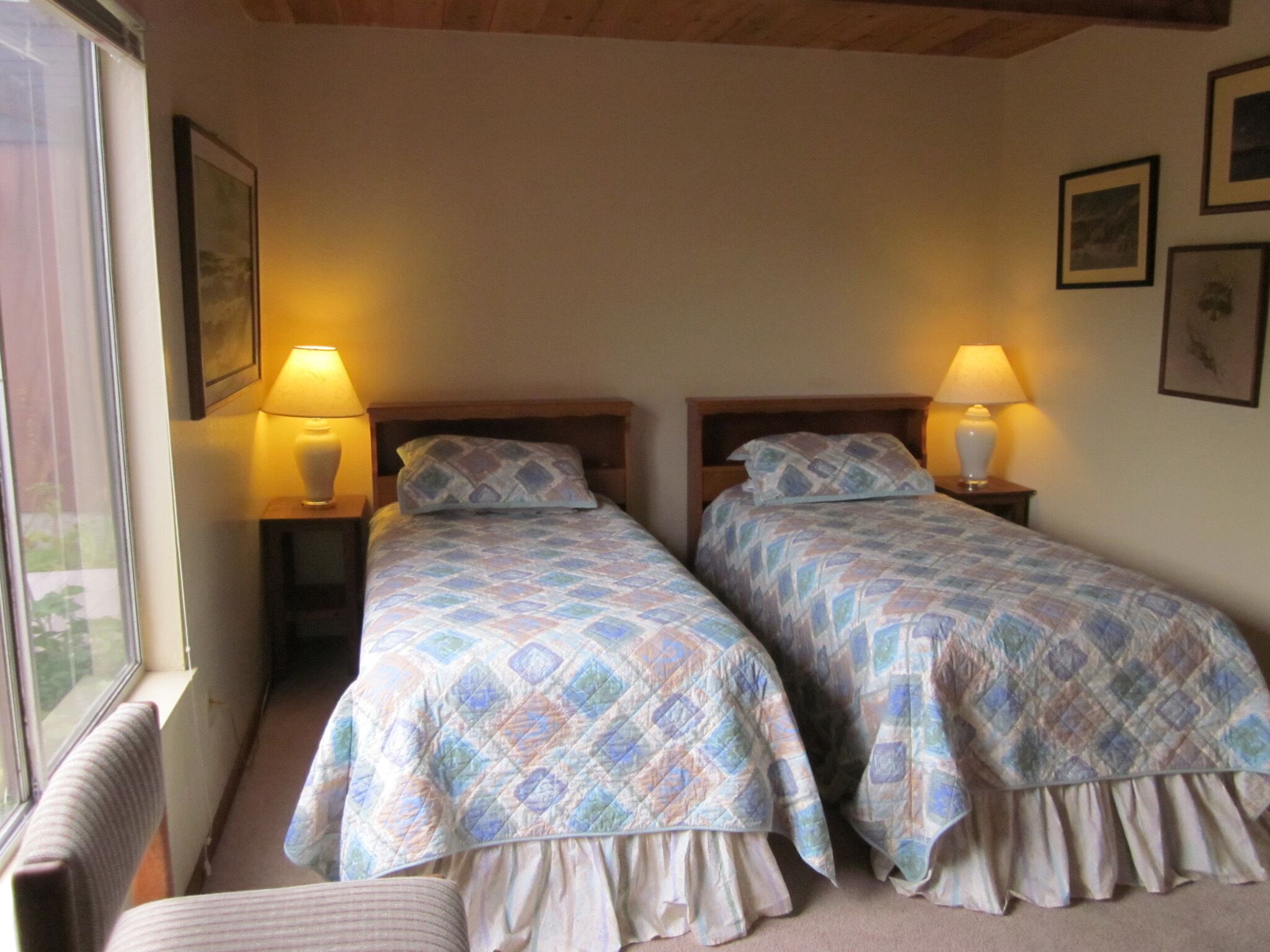 A room with two beds, each with a patterned duvet and a bedside lamp. There is a window on the left side, and a framed picture on the wall.