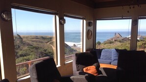 Quarto com grandes janelas que oferecem vista para o oceano e paisagem costeira. Há duas poltronas e uma mesinha com um livro.