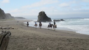Uma praia com pessoas cavalgando, uma grande formação rochosa e o oceano.