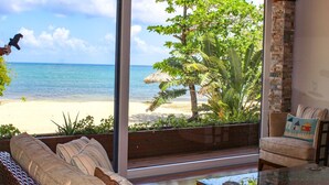 Quarto com uma grande porta de correr de vidro que dá para a praia, um sofá de vime, uma mesa de centro de vidro e uma poltrona acolchoada.